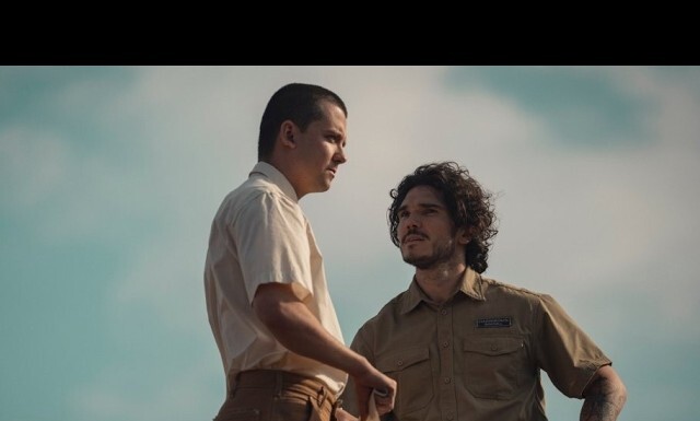 Two men in an outdoor setting stand close, one in a light shirt and the other in a tan uniform, seeming to have a serious conversation.