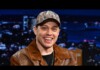Smiling man in a camouflage cap and brown leather jacket on a TV talk show set with city lights backdrop.