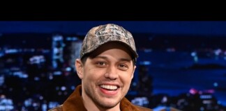 Smiling man in a camouflage cap and brown leather jacket on a TV talk show set with city lights backdrop.
