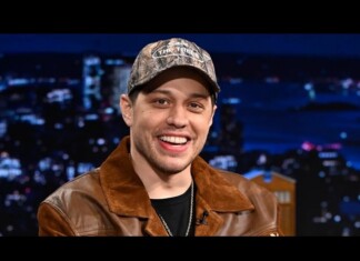 Smiling man in a camouflage cap and brown leather jacket on a TV talk show set with city lights backdrop.