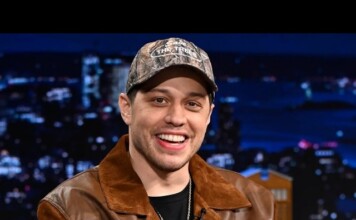 Pete Davidson Reveals Wild Childhood Moment Involving Grandfather and A Gay Porn Video Smiling man in a camouflage cap and brown leather jacket on a TV talk show set with city lights backdrop.