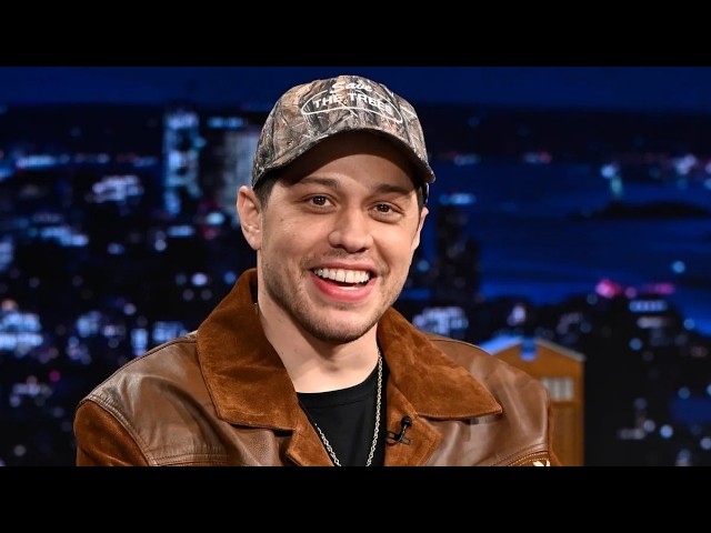 Smiling man in a camouflage cap and brown leather jacket on a TV talk show set with city lights backdrop.