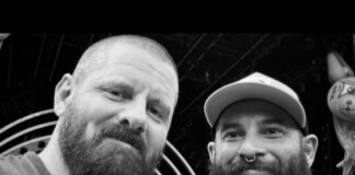 Two bearded men posing for a close-up black-and-white selfie, smiling at the camera, indoors near a circular backdrop.
