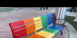 Rainbow-colored metal bench with red to purple slats on a paved plaza area.