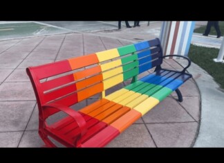 Rainbow-colored metal bench with red to purple slats on a paved plaza area.