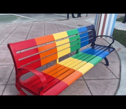 German Tourists Arrested For Nazi Graffiti on Miami Beach Pride Bench Rainbow-colored metal bench with red to purple slats on a paved plaza area.