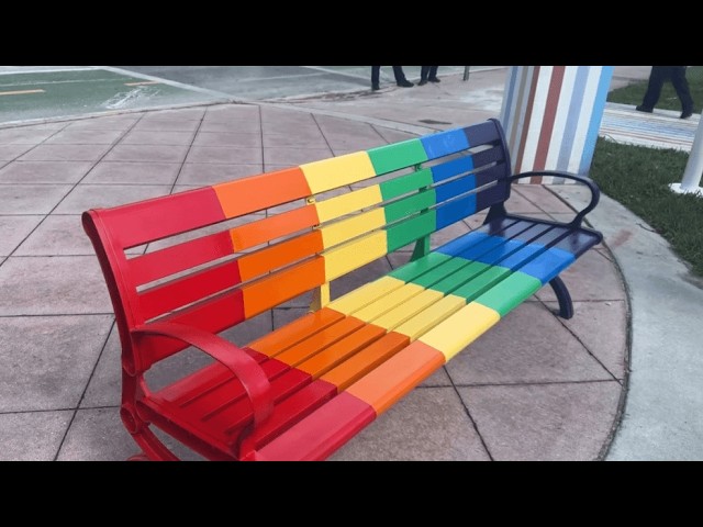 Rainbow-colored metal bench with red to purple slats on a paved plaza area.