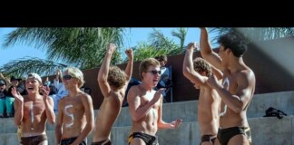 Group of shirtless young male athletes in swim briefs celebrate on concrete bleachers, some with fists raised in victory.
