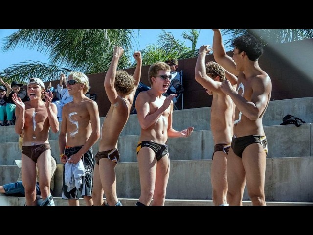 Group of shirtless young male athletes in swim briefs celebrate on concrete bleachers, some with fists raised in victory.