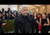 Man with white hair and beard in a black embellished tuxedo on the red carpet as photographers crowd around him.