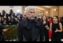 Man with white hair and beard in a black embellished tuxedo on the red carpet as photographers crowd around him.