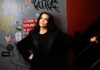 Comedian Jessica Kirson’s Long Road to the Spotlight Finally Pays Off Woman in a black blazer leaning against a graffiti-covered wall near a stair railing, looking up thoughtfully.