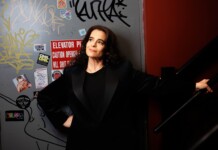 Woman in a black blazer leaning against a graffiti-covered wall near a stair railing, looking up thoughtfully.