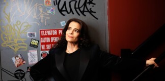 Woman in a black blazer leaning against a graffiti-covered wall near a stair railing, looking up thoughtfully.