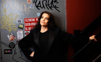 Woman in a black blazer leaning against a graffiti-covered wall near a stair railing, looking up thoughtfully.