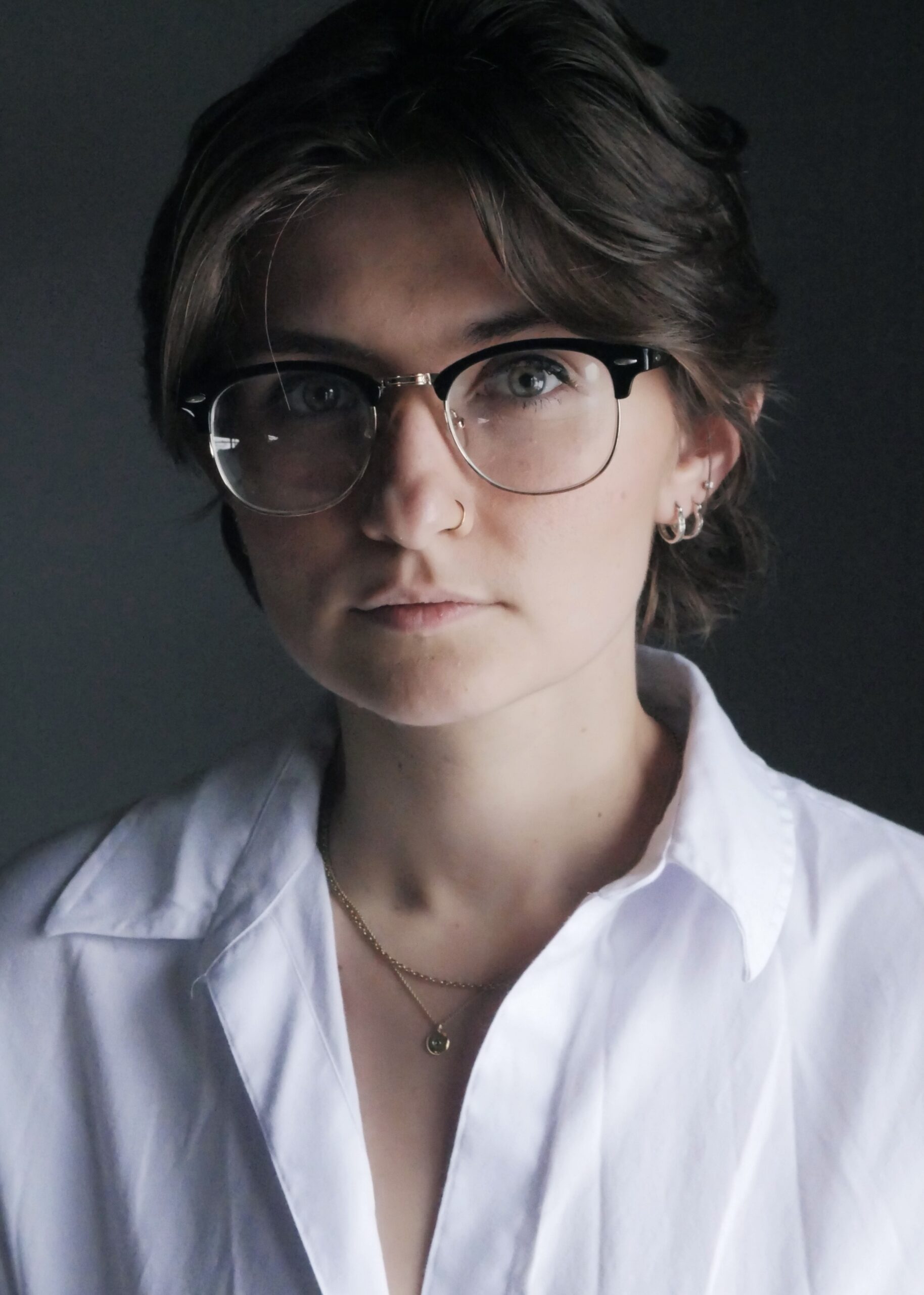 Close-up portrait of a person with short dark hair and round glasses, wearing a white shirt against a dark backdrop.
