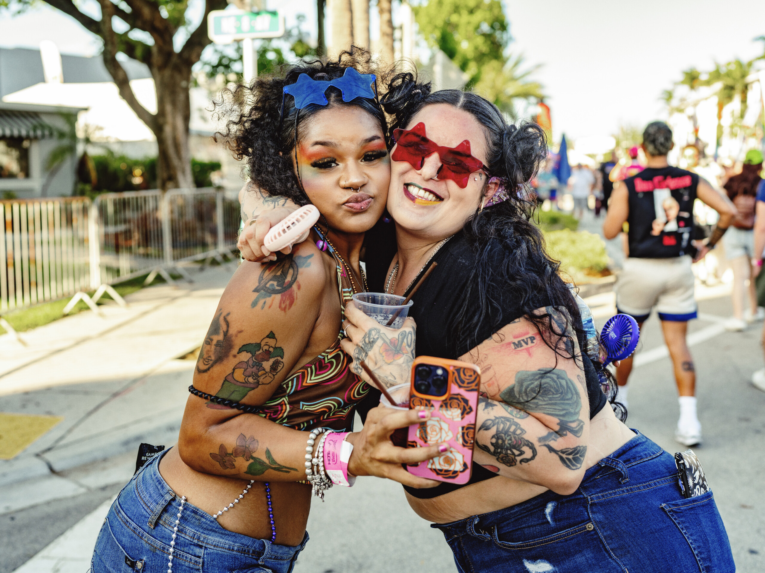 Stonewall Pride Parade and Street Festival Lights up the City of Wilton Manors – Saturday, June 20, 3-11pm. | Hotspots Magazine Hotspots Magazine Two women at a sunny street festival hug for a photo, one wearing bright blue star sunglasses and colorful makeup as they pose.