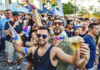 Crowd at a Pride parade cheering and celebrating, with rainbow accessories and drinks in hand, foreground men smiling in sunglasses.