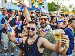 Stonewall Pride Parade and Street Festival Lights up the City of Wilton Manors – Saturday, June 20, 3-11pm. Crowd at a Pride parade cheering and celebrating, with rainbow accessories and drinks in hand, foreground men smiling in sunglasses.