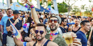 Stonewall Pride Parade and Street Festival Lights up the City of Wilton Manors – Saturday, June 20, 3-11pm. Crowd at a Pride parade cheering and celebrating, with rainbow accessories and drinks in hand, foreground men smiling in sunglasses.