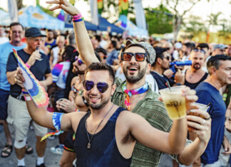Crowd at a Pride parade cheering and celebrating, with rainbow accessories and drinks in hand, foreground men smiling in sunglasses.