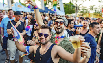 Stonewall Pride Parade and Street Festival Lights up the City of Wilton Manors – Saturday, June 20, 3-11pm. Crowd at a Pride parade cheering and celebrating, with rainbow accessories and drinks in hand, foreground men smiling in sunglasses.