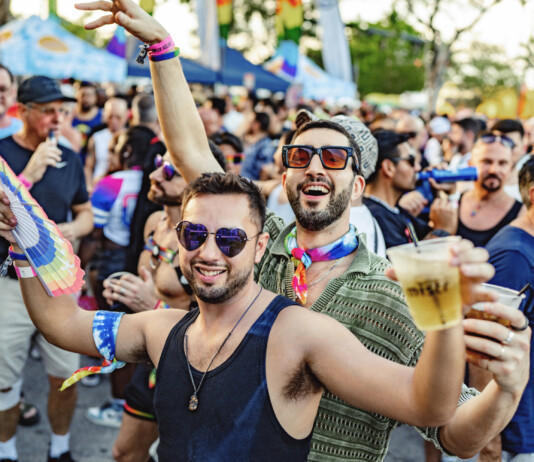 Crowd at a Pride parade cheering and celebrating, with rainbow accessories and drinks in hand, foreground men smiling in sunglasses.