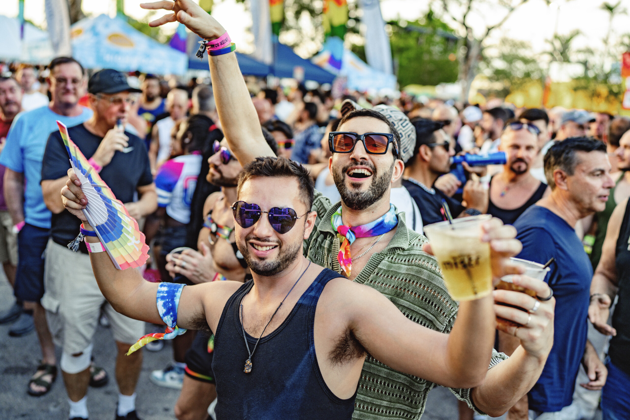 AR500957 Crowd at a Pride parade cheering and celebrating, with rainbow accessories and drinks in hand, foreground men smiling in sunglasses.