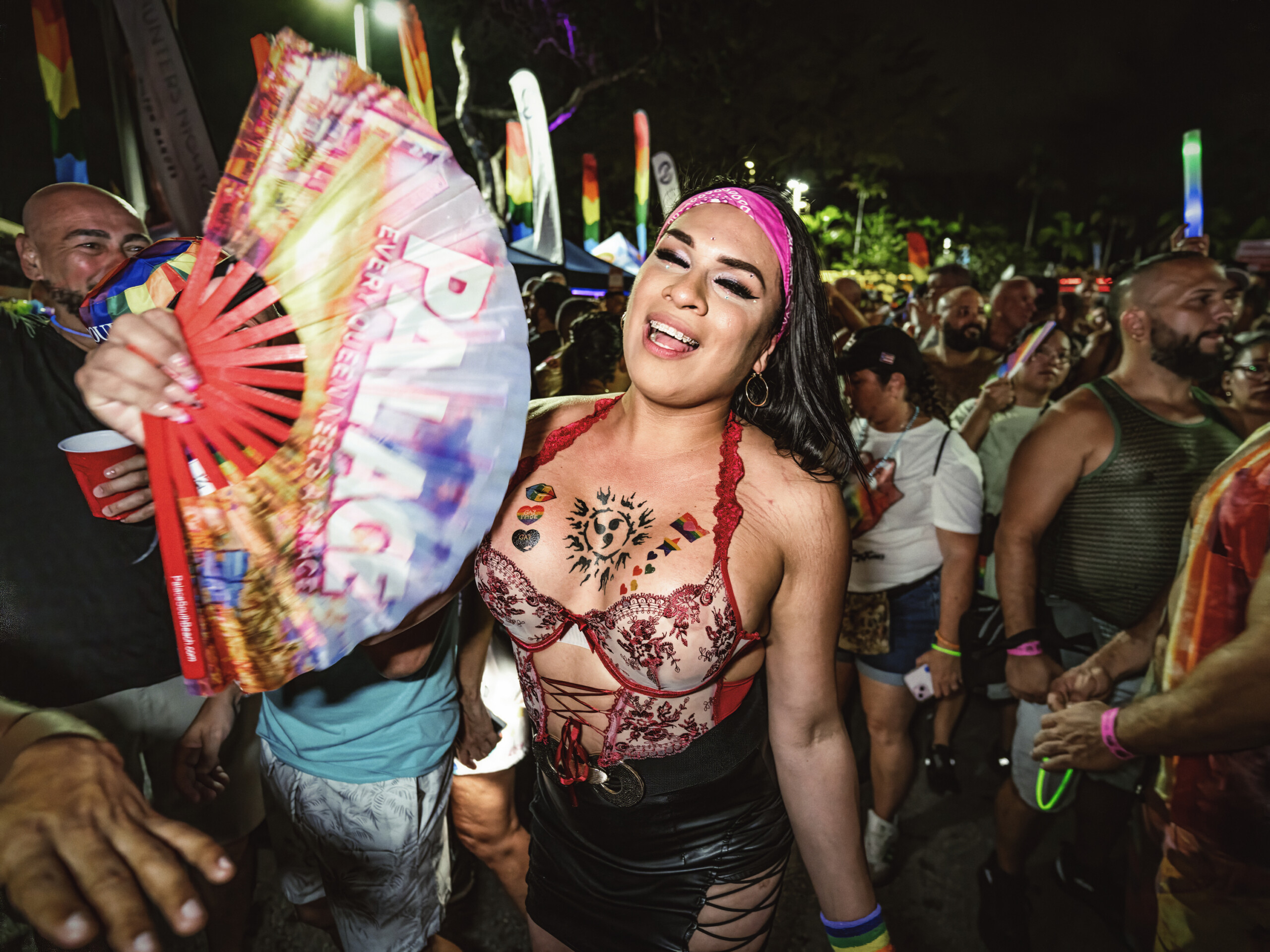 Stonewall Pride Parade and Street Festival Lights up the City of Wilton Manors – Saturday, June 20, 3-11pm. | Hotspots Magazine Hotspots Magazine Joyful Pride festival attendee in a pink headband and red lace top dancing amid a crowd, rainbow flags and colorful fans around at night.