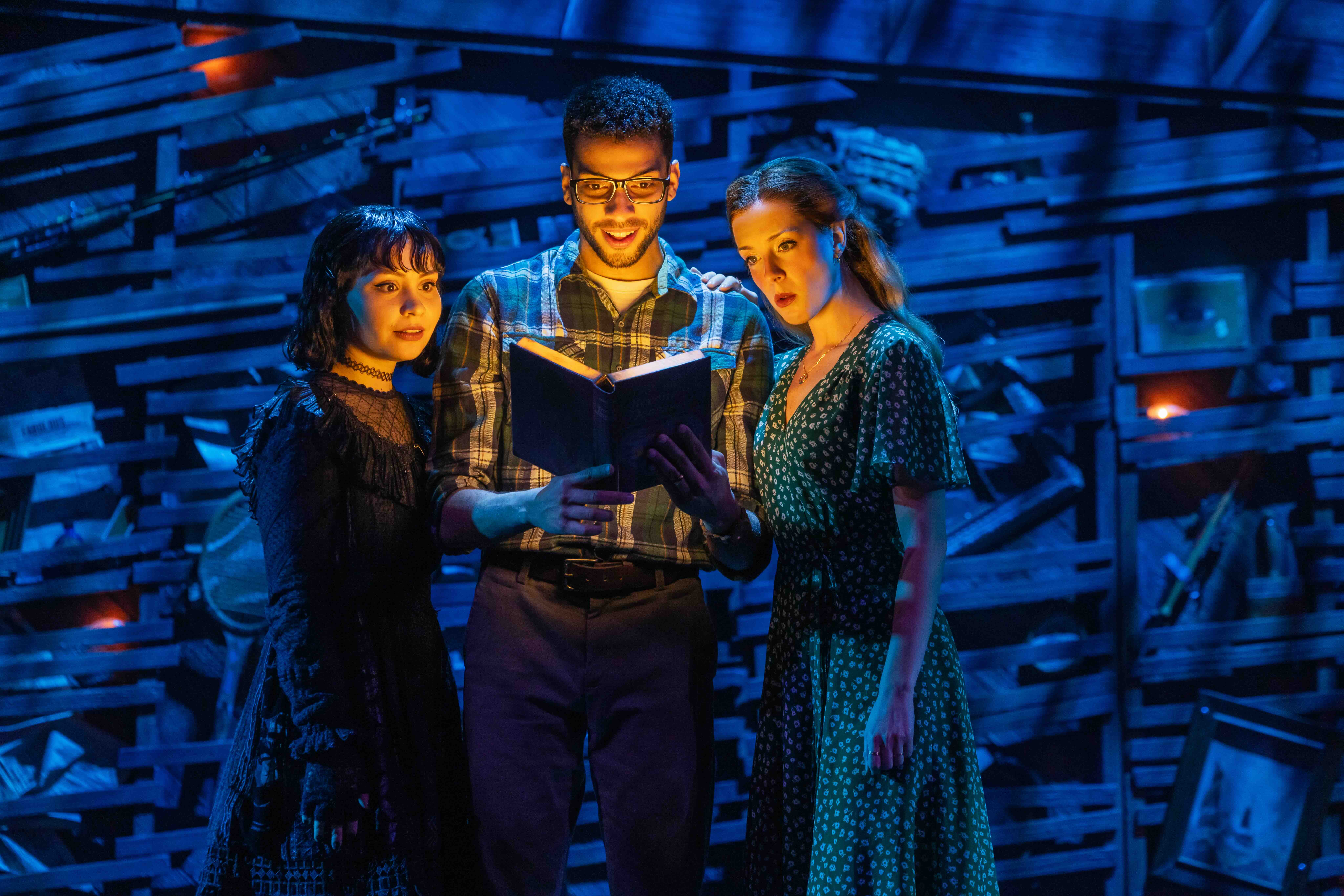 Three people lean in to read an open book together on a blue-lit stage with a rustic pallet backdrop behind them.