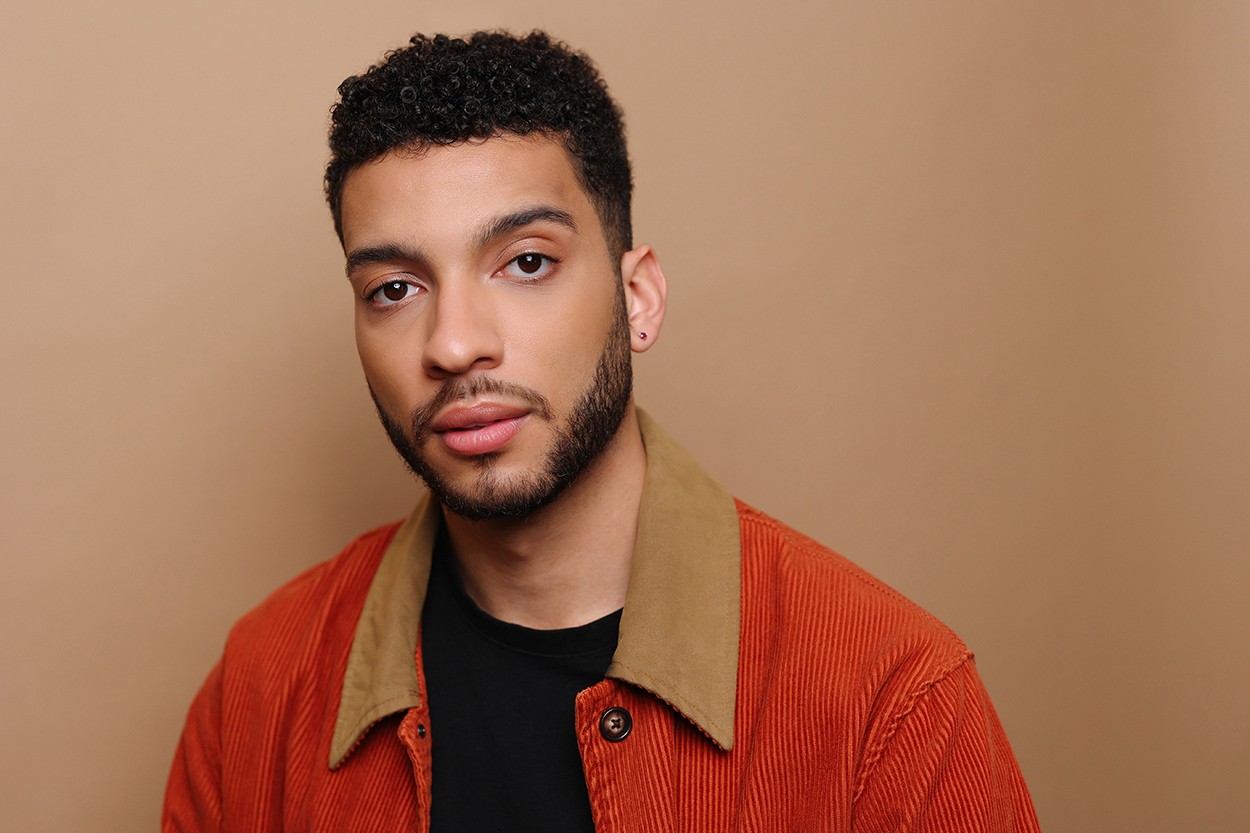 Head-and-shoulders portrait of a man with short curly hair and a beard, wearing an orange corduroy jacket over a black shirt, against a beige backdrop.