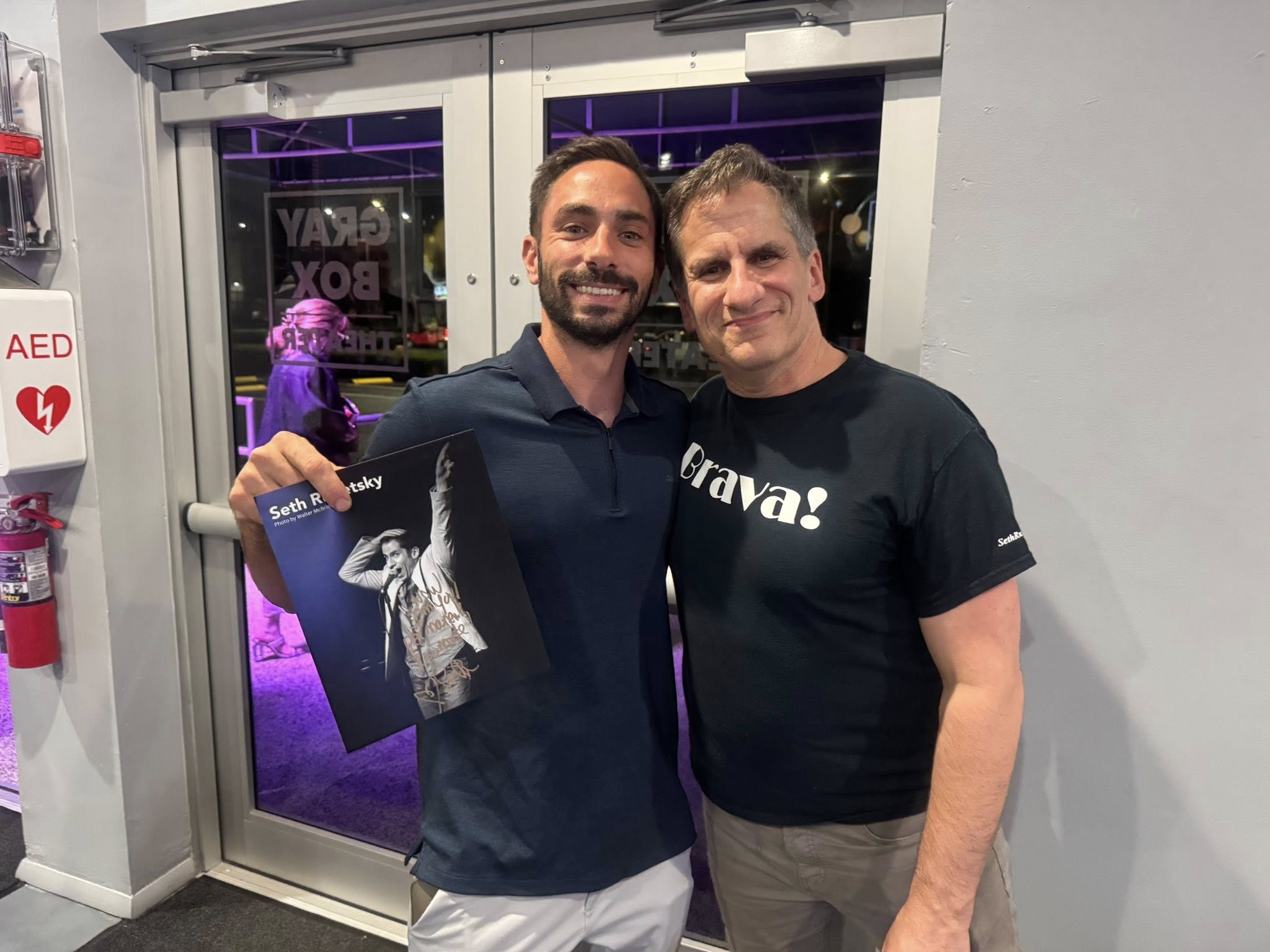 Two men posing for a photo indoors, smiling, standing in front of glass doors with purple lighting in the background.