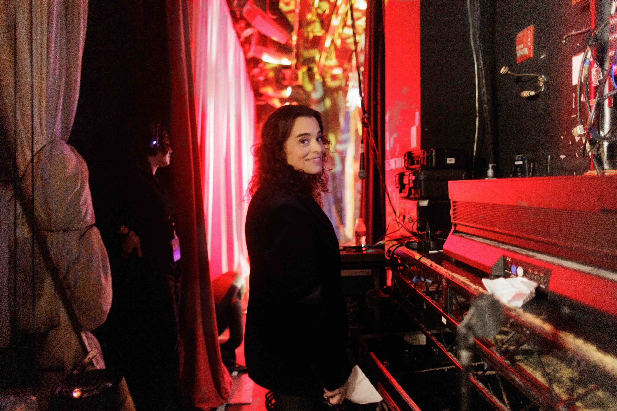 Woman with curly hair smiles while standing beside a red piano/keyboard backstage, with dim red stage lighting and equipment around.