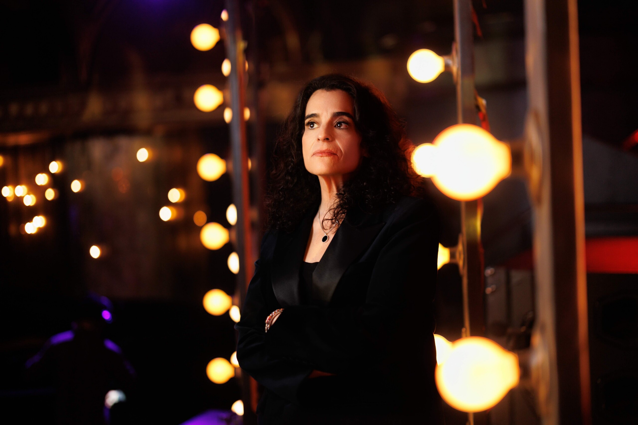 Portrait of a woman with dark, curly hair wearing a black blazer, standing among warm, glowing orbs of light in a dim setting.