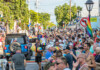 Crowd of people in a sunny Pride parade on a rainbow-filled street with flags and a blue jeep in the foreground.