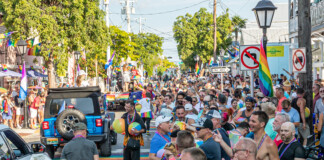Crowd of people in a sunny Pride parade on a rainbow-filled street with flags and a blue jeep in the foreground.