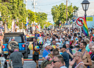 Crowd of people in a sunny Pride parade on a rainbow-filled street with flags and a blue jeep in the foreground.