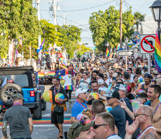 What’s Hot South Florida: June 4 – June 15 Crowd of people in a sunny Pride parade on a rainbow-filled street with flags and a blue jeep in the foreground.