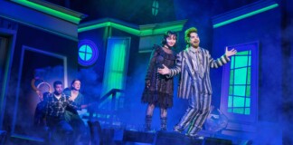 Two actors in vintage costumes headline a blue-lit stage, posing with open arms amid fog.