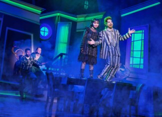 Two actors in vintage costumes headline a blue-lit stage, posing with open arms amid fog.