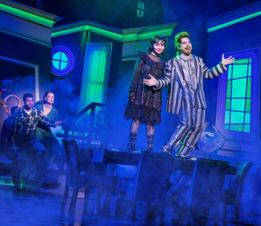 Two actors in vintage costumes headline a blue-lit stage, posing with open arms amid fog.