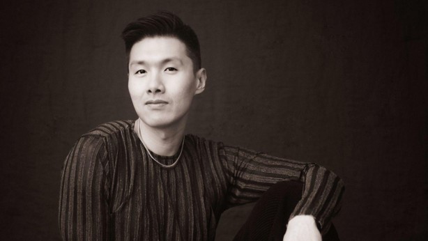Portrait of a young man with short dark hair wearing a striped knit sweater, seated against a dark background.
