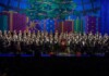 Gay Men’s Chorus of South Florida Brings Arts Groups Together Large choir with an orchestra on a festively decorated stage, Christmas tree and colorful presents in the background, audience in silhouette front rows