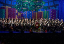 Large choir with an orchestra on a festively decorated stage, Christmas tree and colorful presents in the background, audience in silhouette front rows