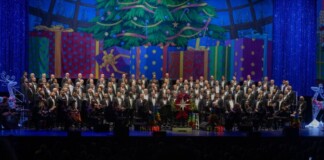 Large choir with an orchestra on a festively decorated stage, Christmas tree and colorful presents in the background, audience in silhouette front rows