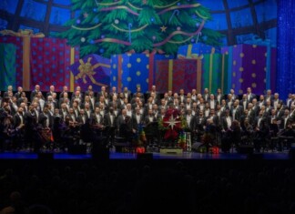Gay Men’s Chorus of South Florida Brings Arts Groups Together Large choir with an orchestra on a festively decorated stage, Christmas tree and colorful presents in the background, audience in silhouette front rows
