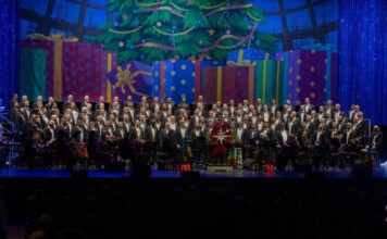 Large choir with an orchestra on a festively decorated stage, Christmas tree and colorful presents in the background, audience in silhouette front rows