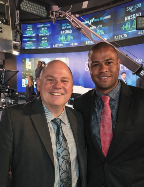 Two men in business suits smile for a photo on a stock trading floor, with large market ticker screens behind them.