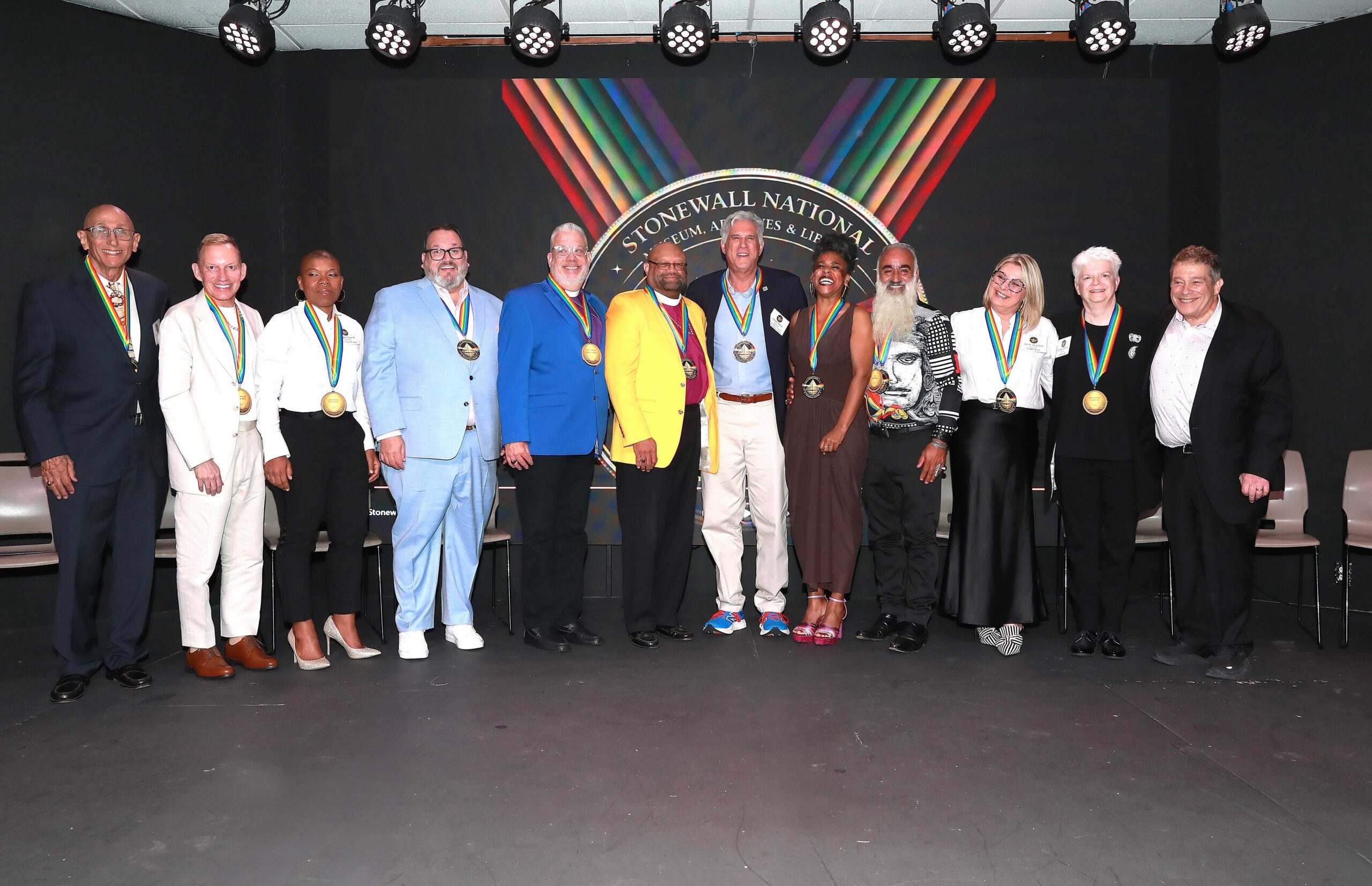 Group of diverse adults in formal attire wearing medals, posing on a stage in front of a Stonewall National backdrop.