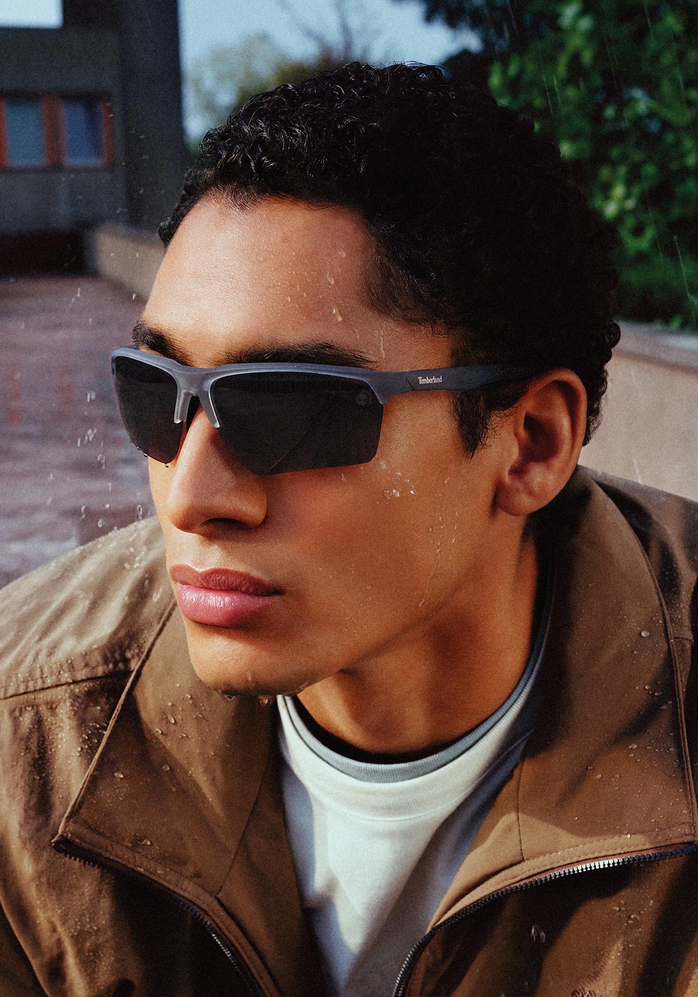 Person wearing dark sunglasses and a brown jacket in the rain, close-up portrait with droplets on glass or lens nearby, Timberland logo visible on sunglasses.